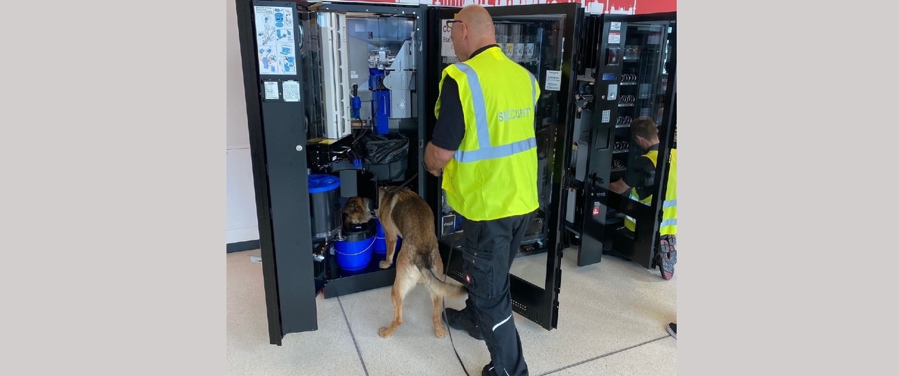 Hund und Hundeführer beschnüffeln einen Getränkeautomaten:  Sie machen den BER sauber.