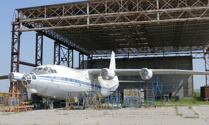 Verlassen steht eine Antonov An-12 vor einem ehemaligen Hangar. Beide rosten vor sich hin.
