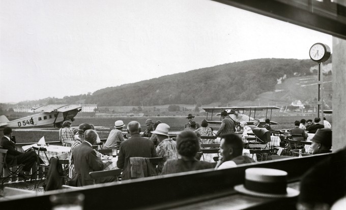 Gut besucht ist im Sommer 1934 die Terrasse des Flugplatz-Restaurants auf dem Sternenfeld. Auf dem Flug der Deutschen Luft Hansa nach Frankfurt wird die Dornier Merkur D 546 eingesetzt. Die D 546 wurde 1925 als Dornier Komet III gebaut und «Hyäne» genannt. Betrieben wurde sie durch die damals grösste deutsche Fluggesellschaft Aero Lloyd. Am 1. Januar 1926 wurde sie bei der Gründung der Deutschen Luft Hansa in die Gesellschaft eingebracht. Die Luft Hansa liess die an schwacher Motorleistung krankende Komet III zum Nachfolgemuster Merkur umbauen, u.a. erkennbar am modifizierten Leitwerk und am unterhalb des Motors angebrachten Kühler. Rechts im Bild ist die Caudron C.59 CH-125 geparkt, vermutlich die einzige in der Schweiz registrierte Maschine dieses Typs, von dem aber in Frankreich rund 1'800 Einheiten gebaut wurden.