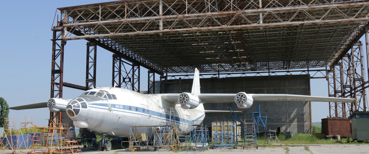 Verlassen steht eine Antonov An-12 vor einem ehemaligen Hangar. Beide rosten vor sich hin.